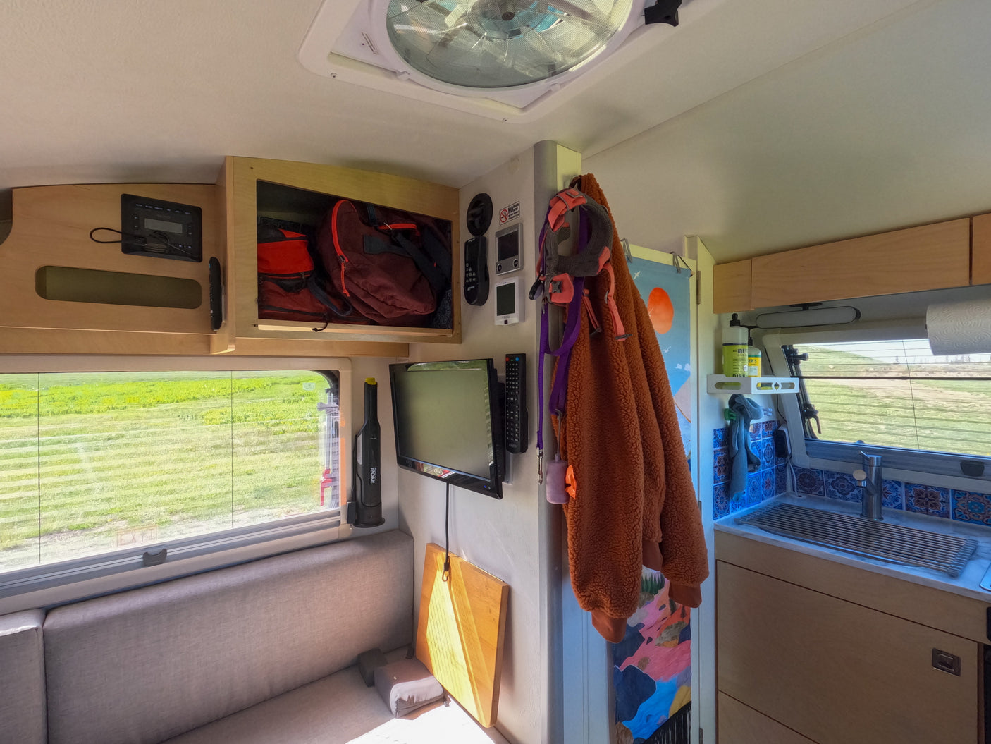 Bright RV rental interior, featuring a sofa, window with a view, TV, overhead storage, and a tiled kitchenette.