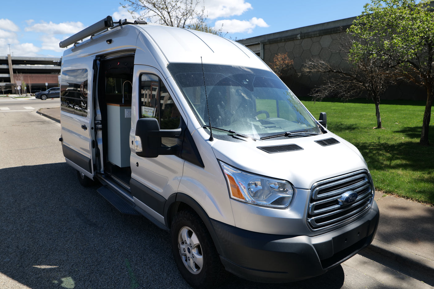 Silver Ford Transit camper van for rv rental, showing open side door with custom interior, parked outdoors on a sunny day.