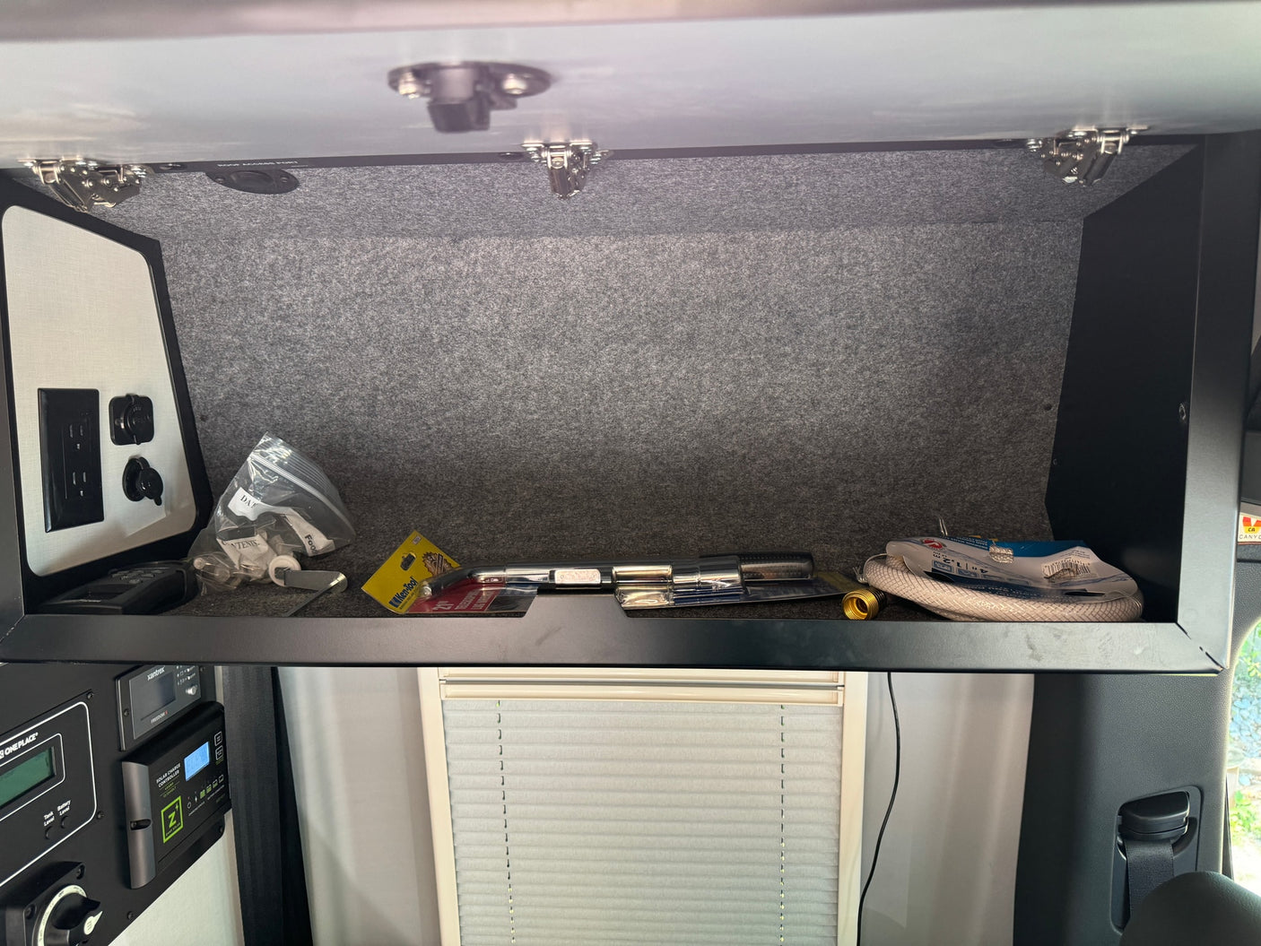 An RV rental's overhead cabinet displaying tools, hoses, and an electrical control panel with a solar charge controller.