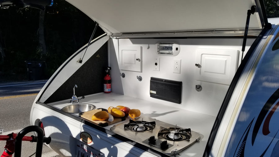 Open rear kitchen of an RV rental teardrop camper, featuring a sink, stove, fruit on counter, and safety equipment.