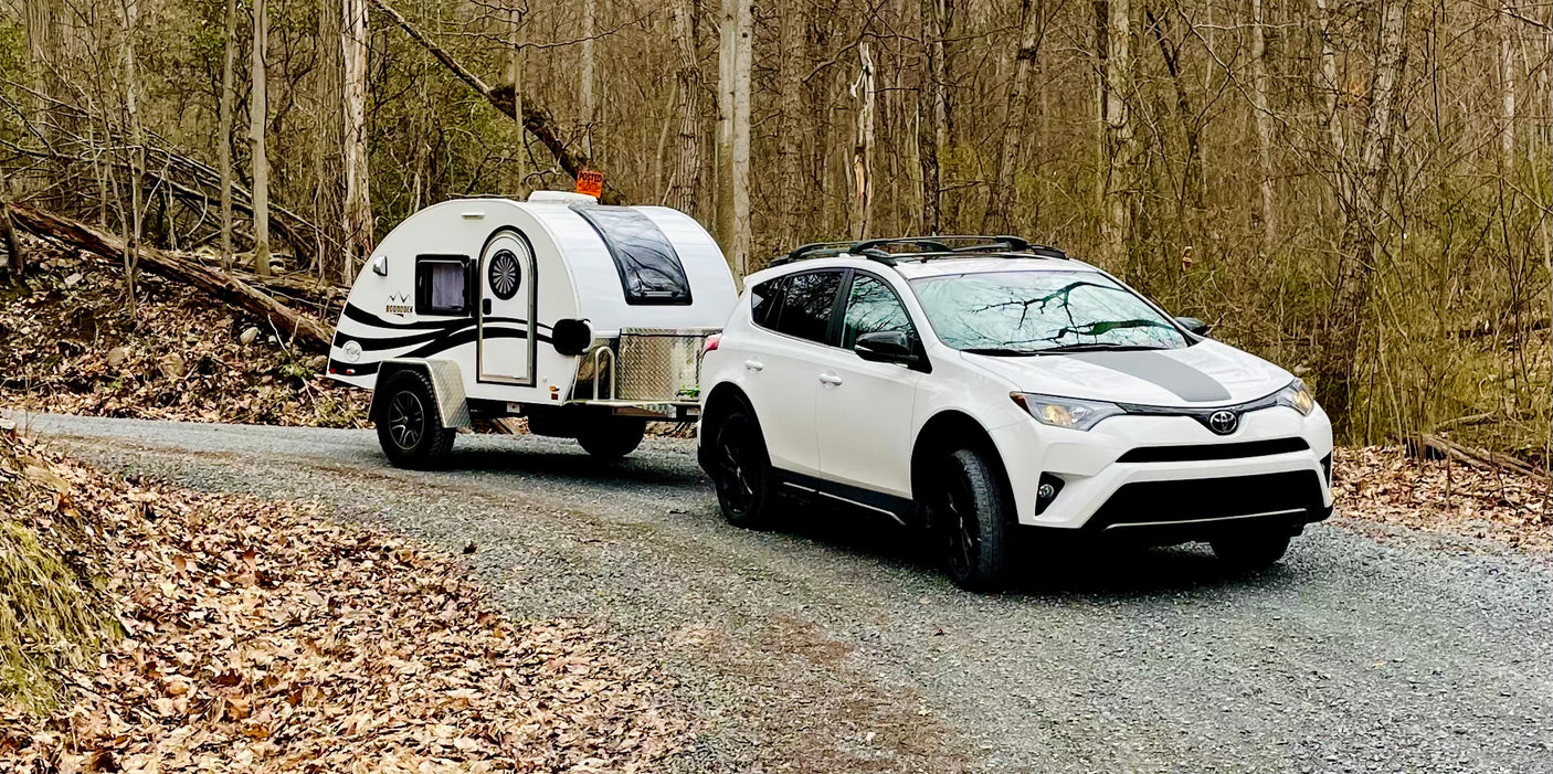White SUV towing a teardrop RV rental on a gravel road through a bare forest. Ready for outdoor adventure.