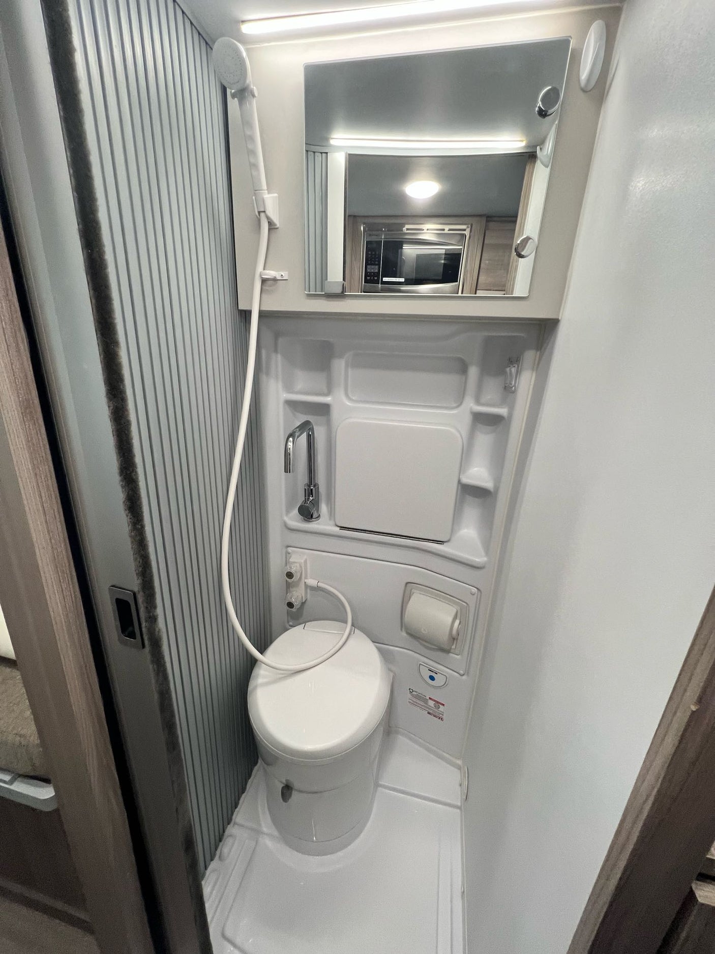 Compact RV rental bathroom showing toilet, shower, sink, and mirror reflecting a microwave.