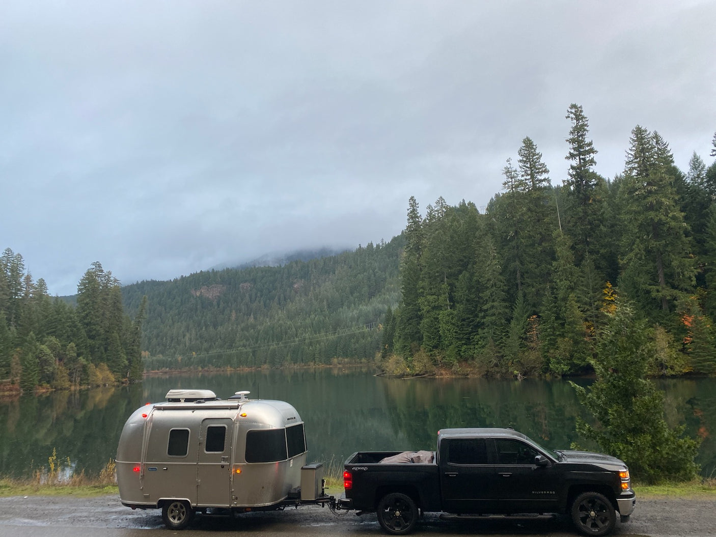 RV rental: A silver Airstream trailer hitched to a truck, parked by a calm lake with forested, misty mountains.