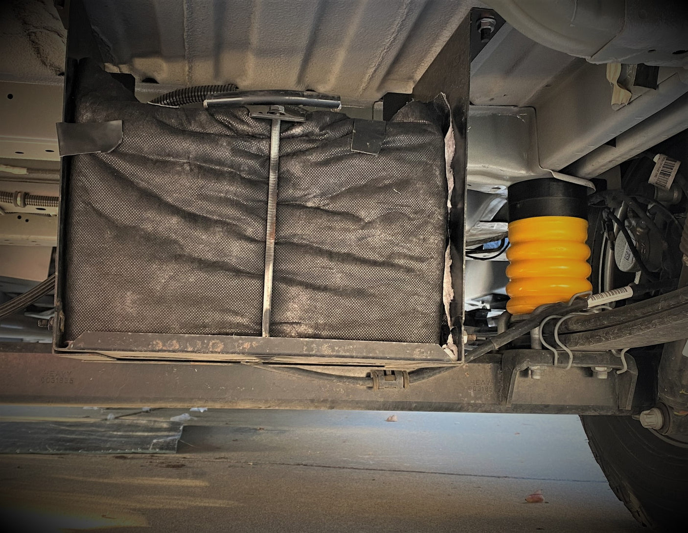 Underside of an RV rental showing a mounted battery box and a bright yellow suspension helper spring.