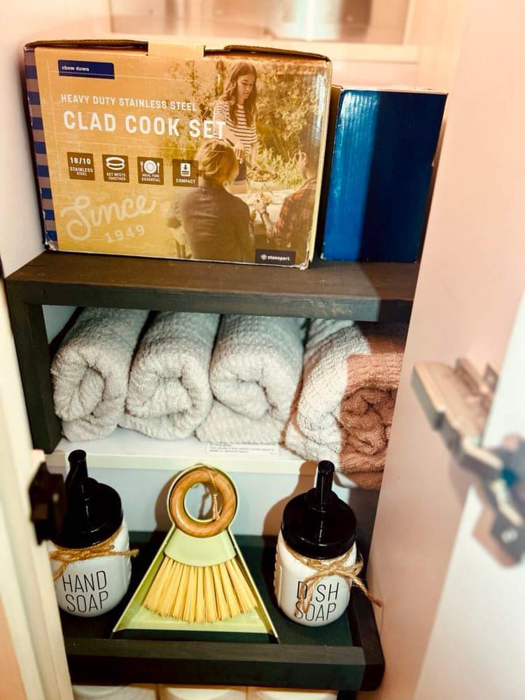 Compact Clad Cook Set, rolled towels, hand/dish soap, and cleaning brush stored in neat shelves for an RV rental.