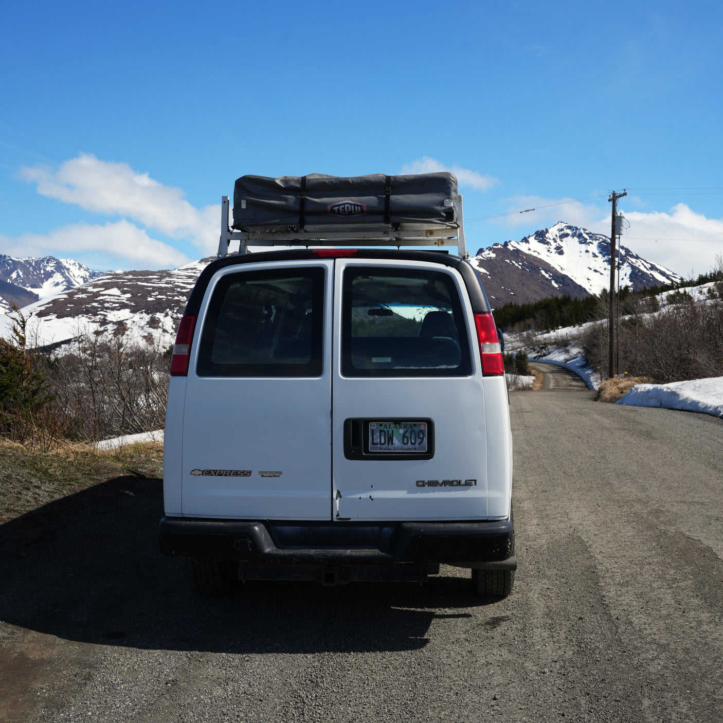 White cargo van RV rental with rooftop tent on a scenic gravel road in snowy Alaskan mountains under blue sky.