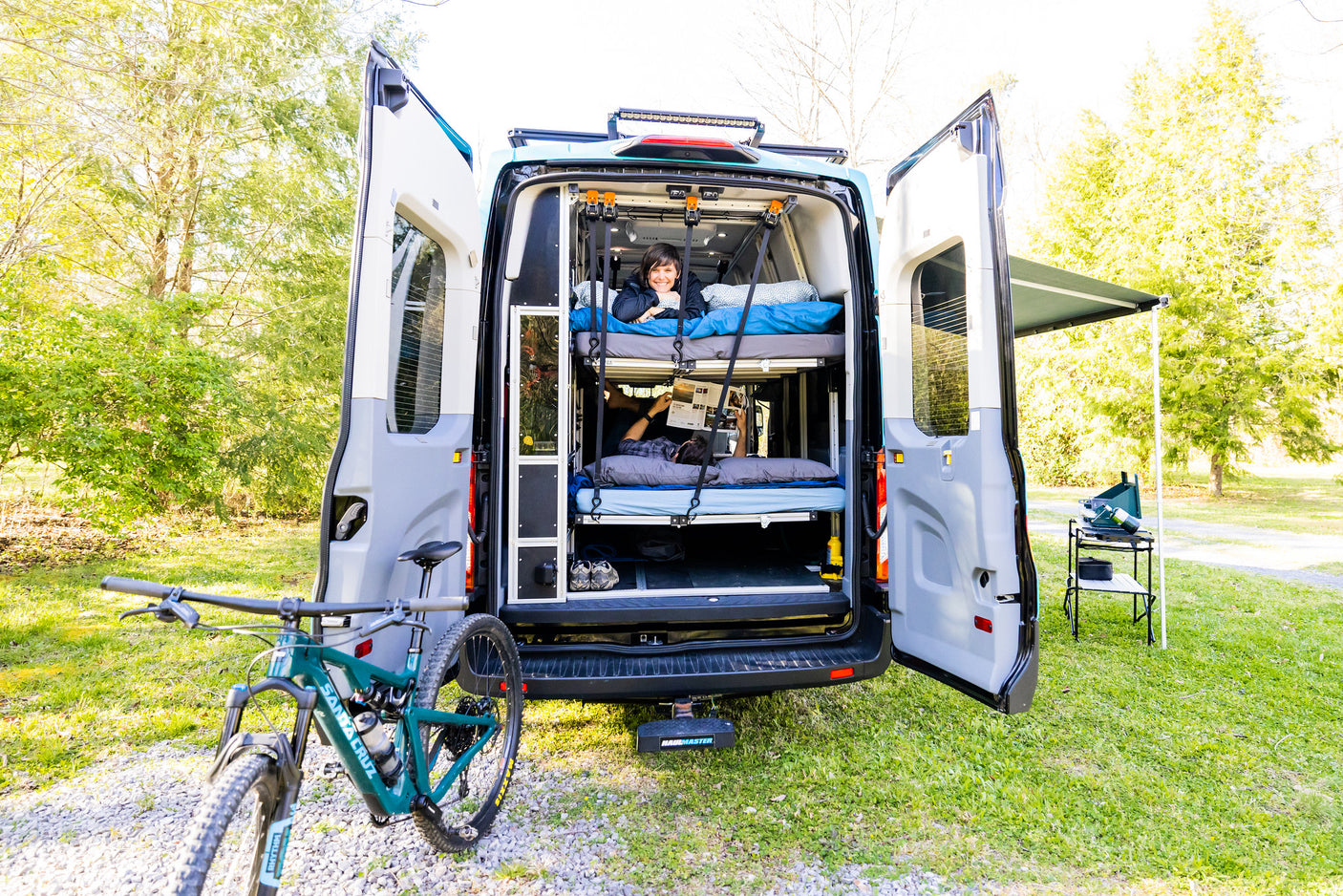 Open RV rental camper van reveals bunk beds with people relaxing, plus a mountain bike & outdoor cooking setup.