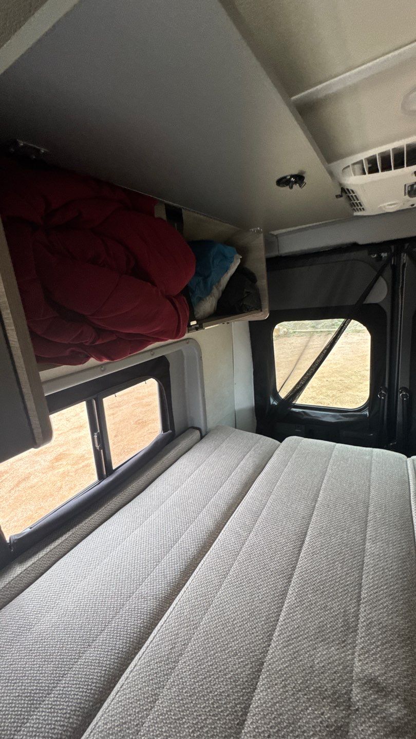 RV rental interior: cozy bed, overhead storage with bedding, and side windows revealing an outdoor view. Ideal for travel.