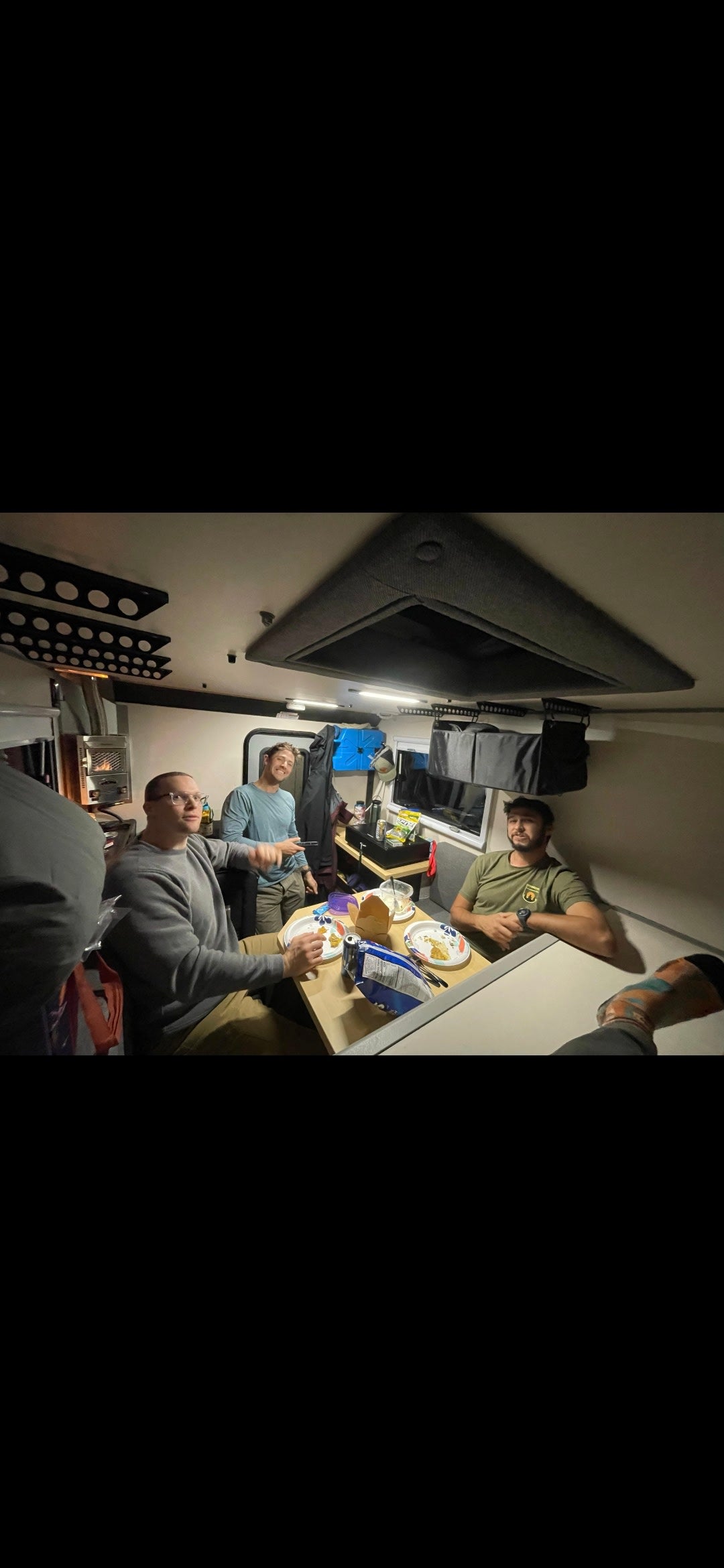 Four men share a meal at a table inside a compact, well-lit rv rental camper, featuring a heater. Perfect for travel adventures.