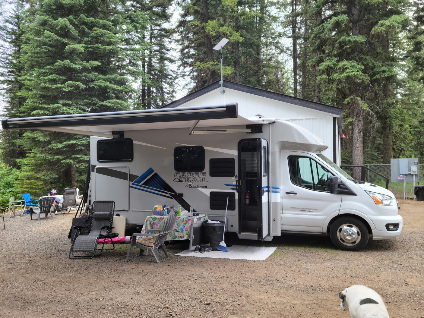 White RV rental at a wooded campsite, awning extended over outdoor dining area with chairs & table. Dog on gravel foreground.