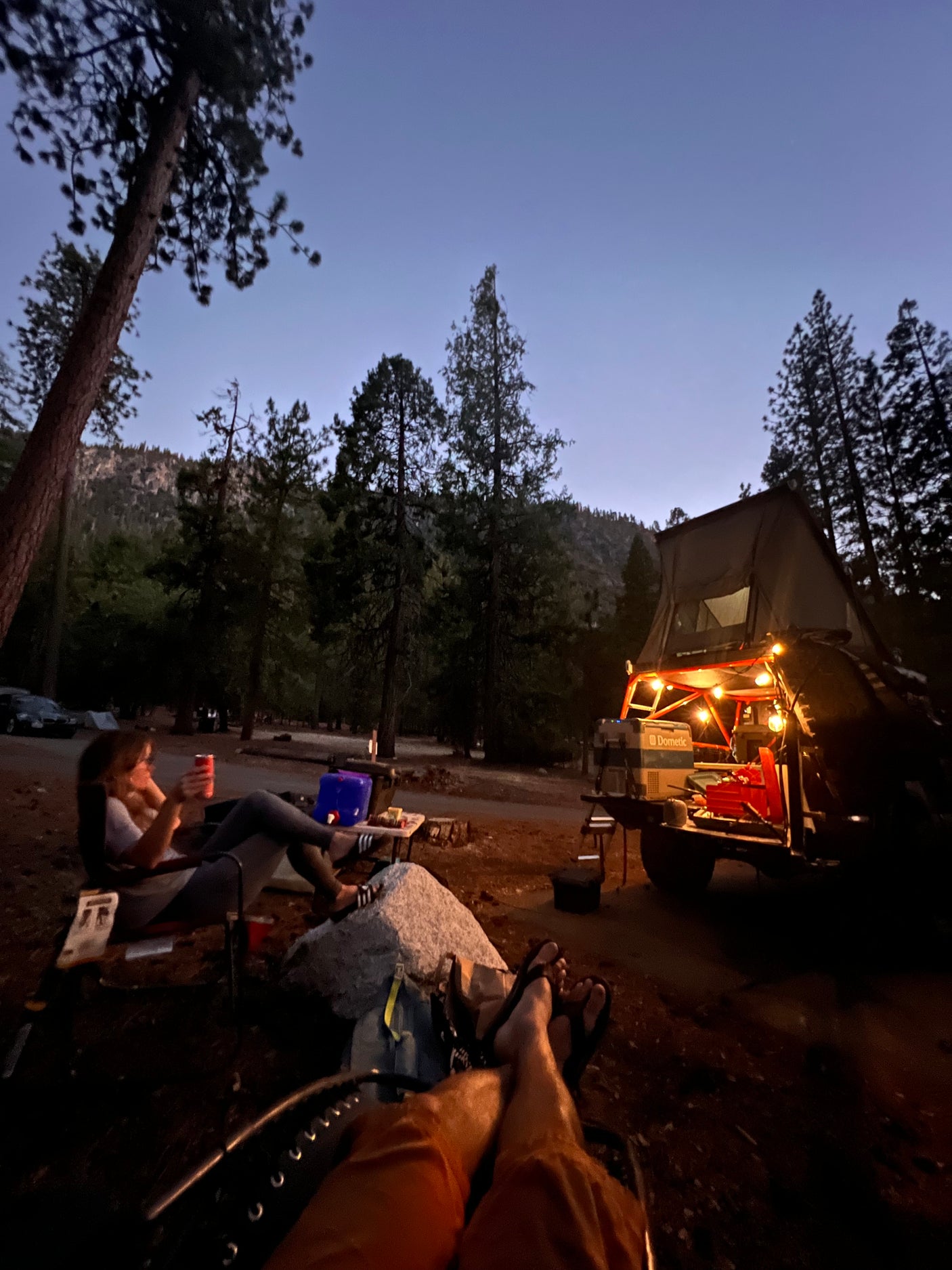 Twilight camping scene: People relaxing at an RV rental site. Rooftop tent, vehicle, and festive lights illuminating the forest campsite.