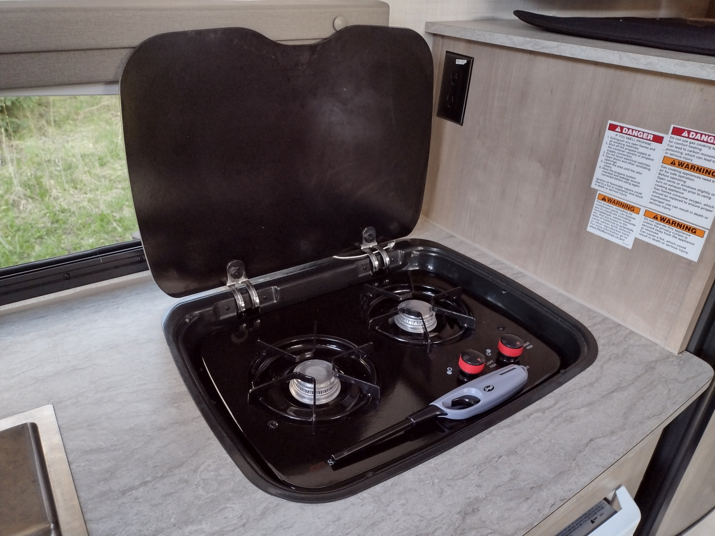 RV rental kitchen showing an open two-burner gas stove, a lighter, a sink, and essential safety labels on the wall.