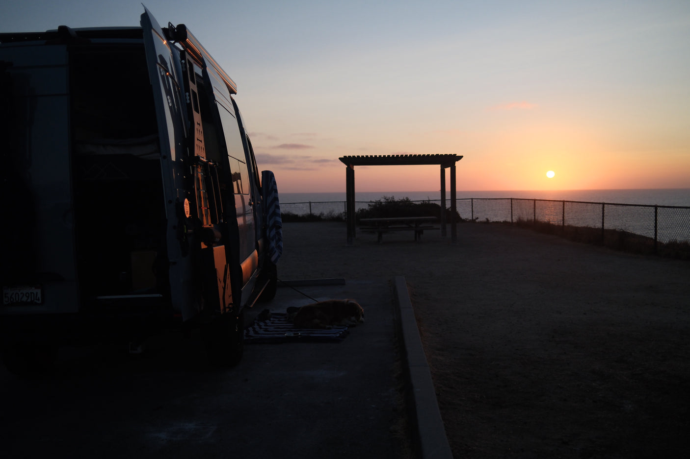 RV rental sunset: A van, dog, and pergola overlook a serene ocean view at dusk. Peaceful camping.