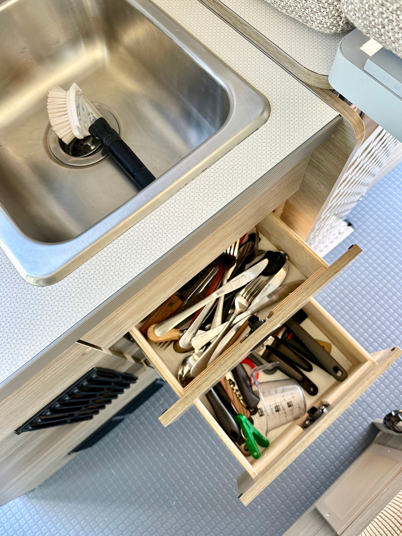 Inside an RV rental: a compact kitchen features a stainless steel sink with brush and open wooden drawers neatly holding cutlery and tools.