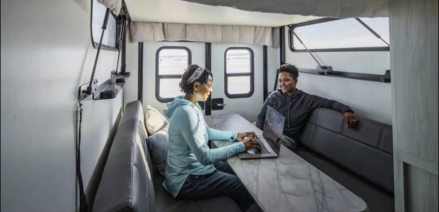 Spacious RV rental interior: two women, one on laptop at table, other relaxing, with scenic outdoor views from windows.
