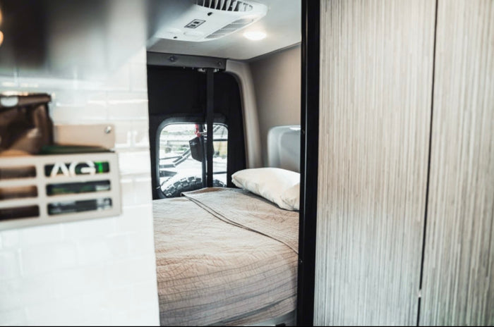 Cozy rv rental interior: a comfortable bed with a quilted blanket and pillow, and an AC unit in a compact camper van.