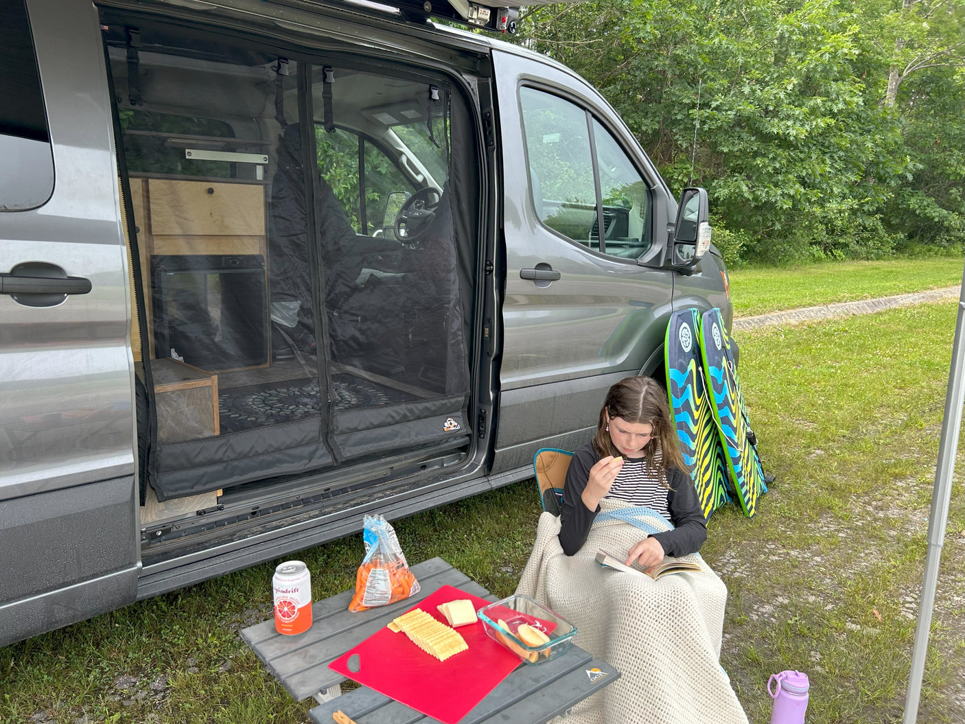 Child reads by a grey rv rental van, with snacks on a table and boogie boards outside for adventures.