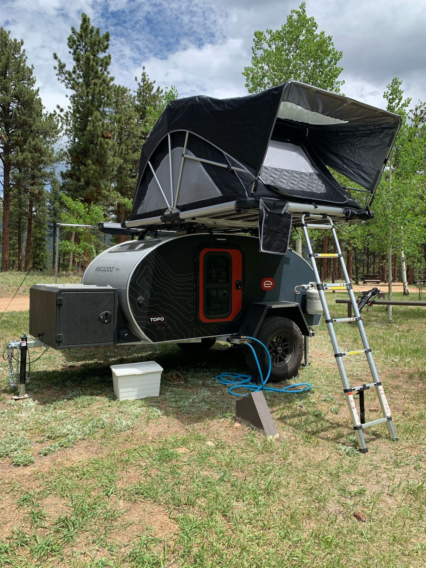 An Escapod topo teardrop trailer with a rooftop tent set up for an rv rental camping adventure in a forest.