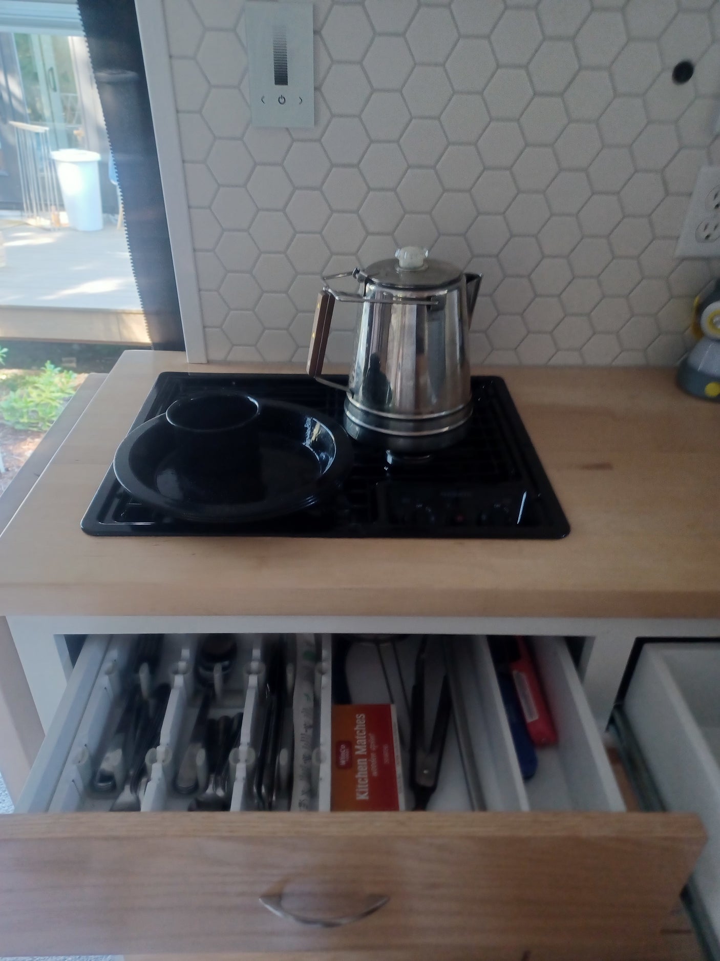 RV rental kitchen: stovetop with percolator, hexagonal backsplash, and an open drawer with organized cutlery and matches.