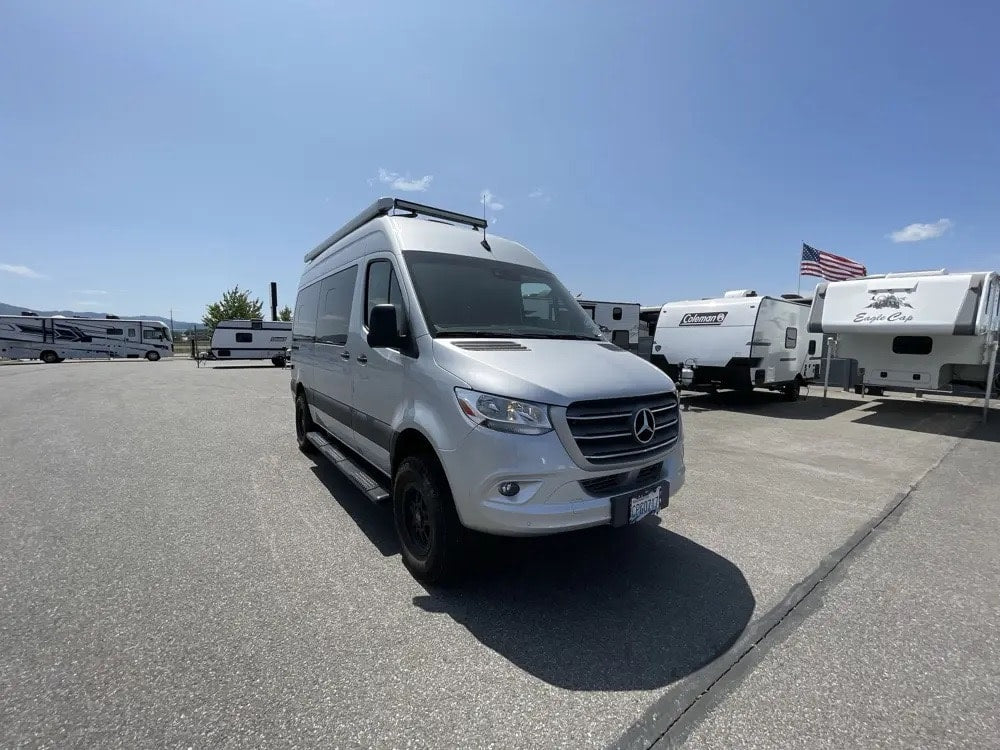 parked on gravel with trees & an Airstream trailer.|Side view of a silver Mercedes-Benz Sprinter "Tranquility" camper van