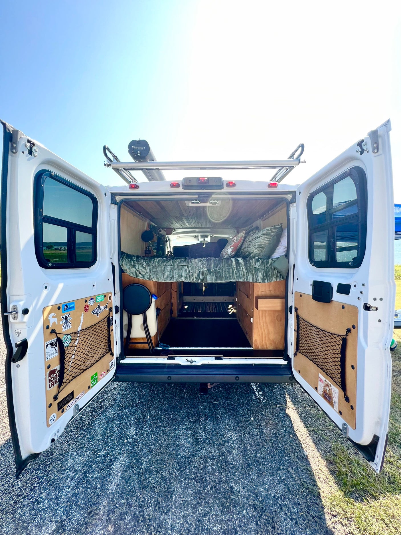Open rear doors of this custom rv rental van show a wood interior, bed, storage, and a Road Shower 4 on the roof.