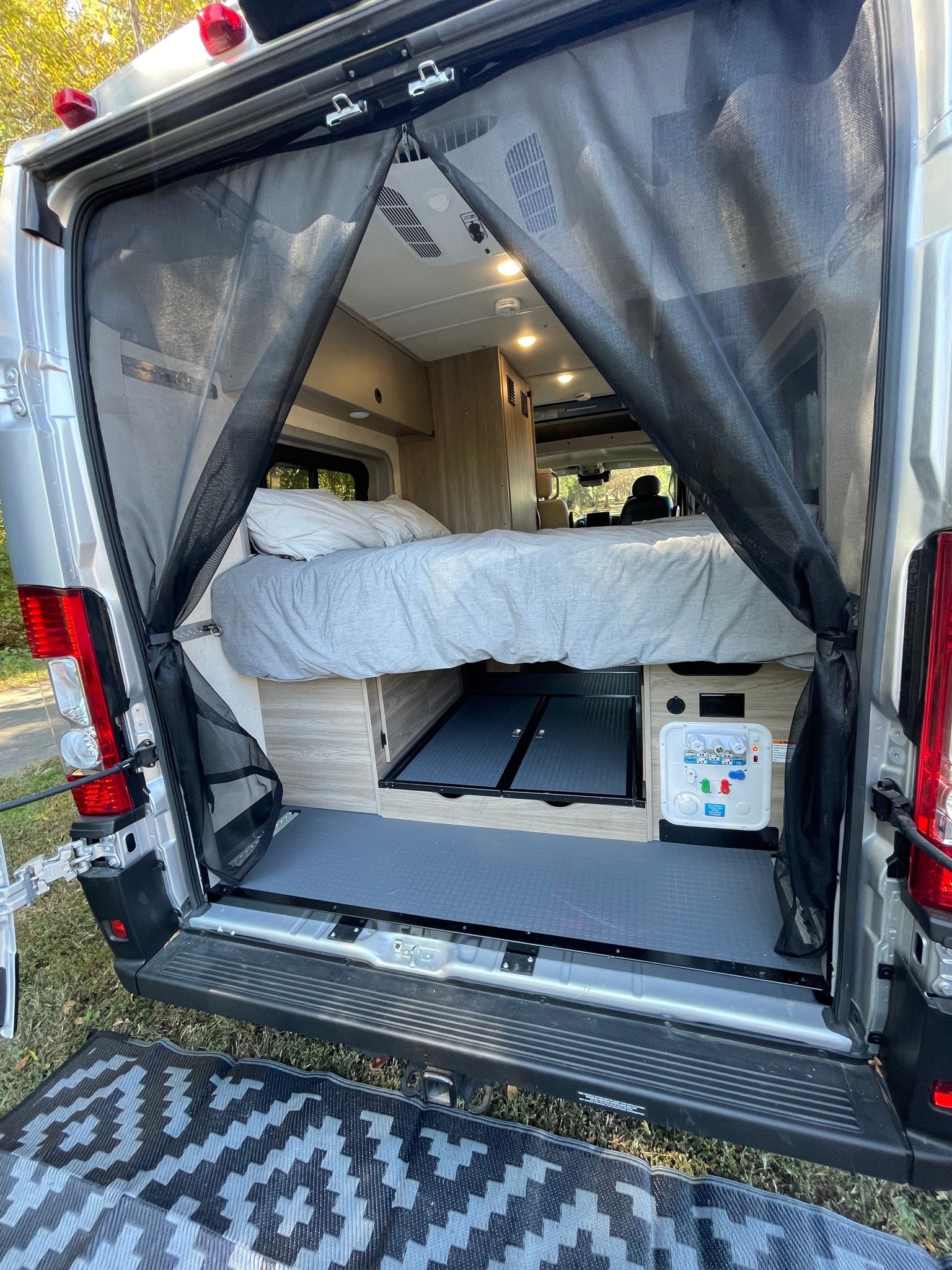 RV rental: Rear view of a camper van, showcasing its bed, interior storage, and a magnetic screen door. Outdoor mat visible.