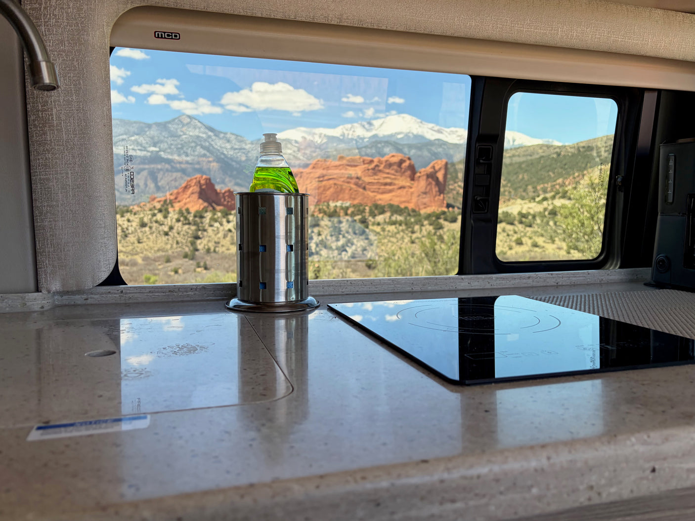 Inside a modern rv rental: kitchen counter, induction cooktop, dish soap. Outside, red rock formations & snowy mountains.