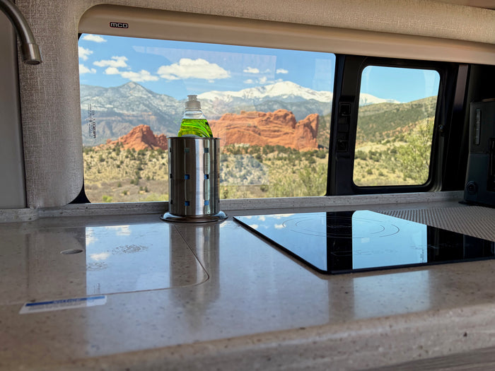 Inside a modern rv rental: kitchen counter, induction cooktop, dish soap. Outside, red rock formations & snowy mountains.