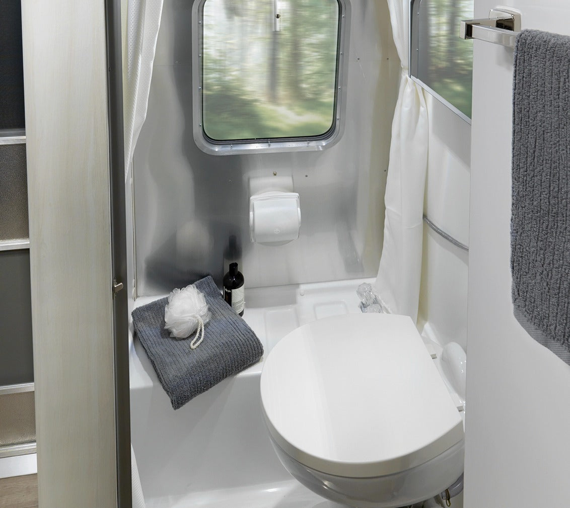 Inside an RV rental: a compact bathroom with a toilet, shower curtain, and window showing blurred trees. Includes a towel and toiletries.