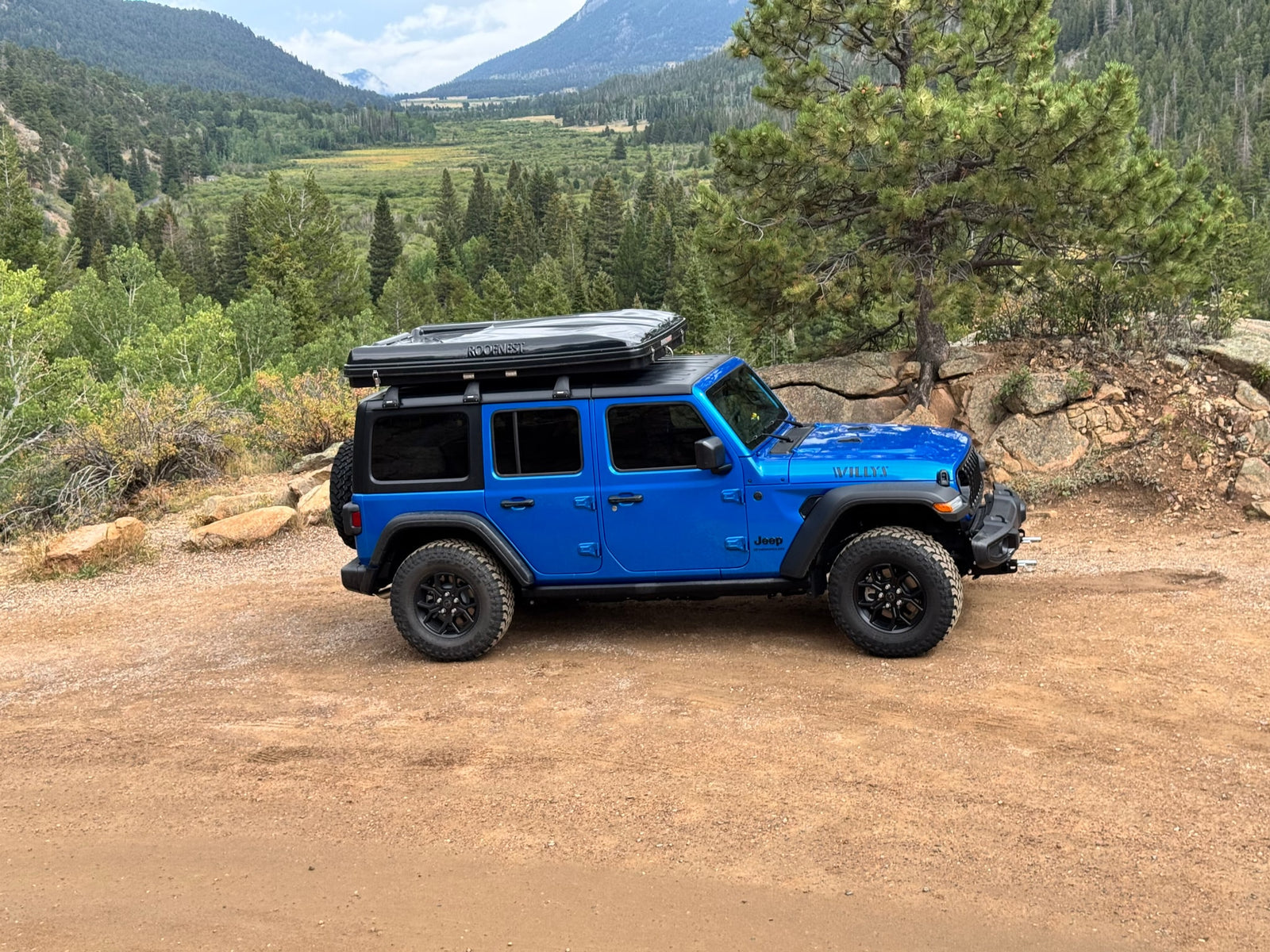 A vibrant blue Jeep Wrangler with a Roofnest rooftop tent on a dirt road, amidst lush mountains. Ideal for an RV rental adventure.