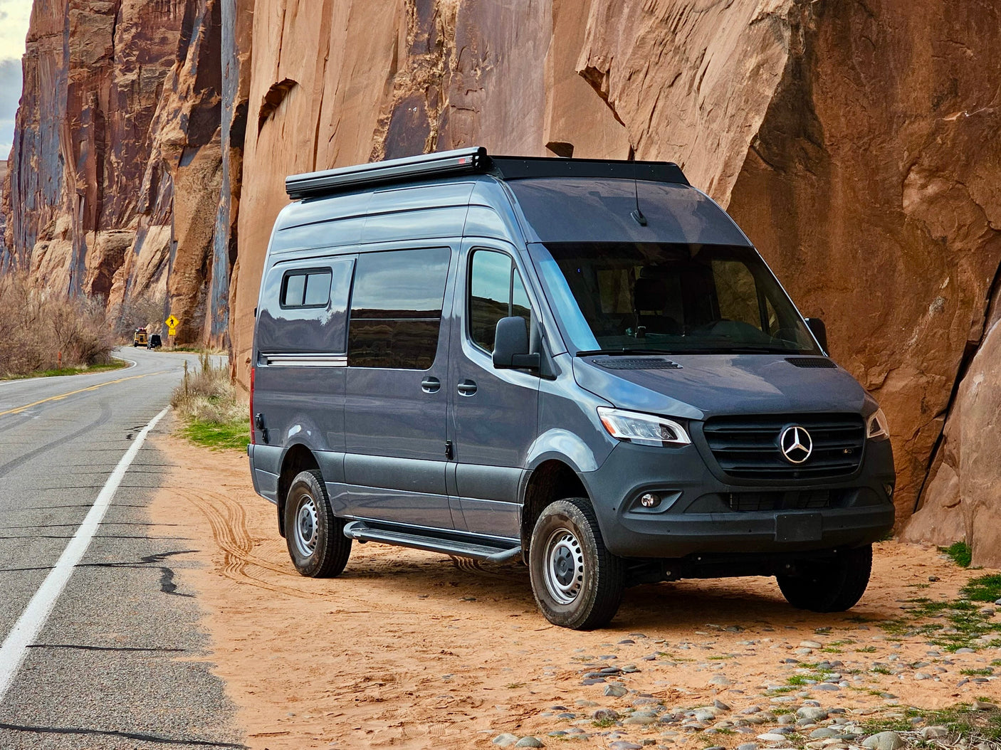 Dark grey Mercedes-Benz Sprinter camper van (RV rental) parked on a dirt road shoulder beside towering red rock cliffs.