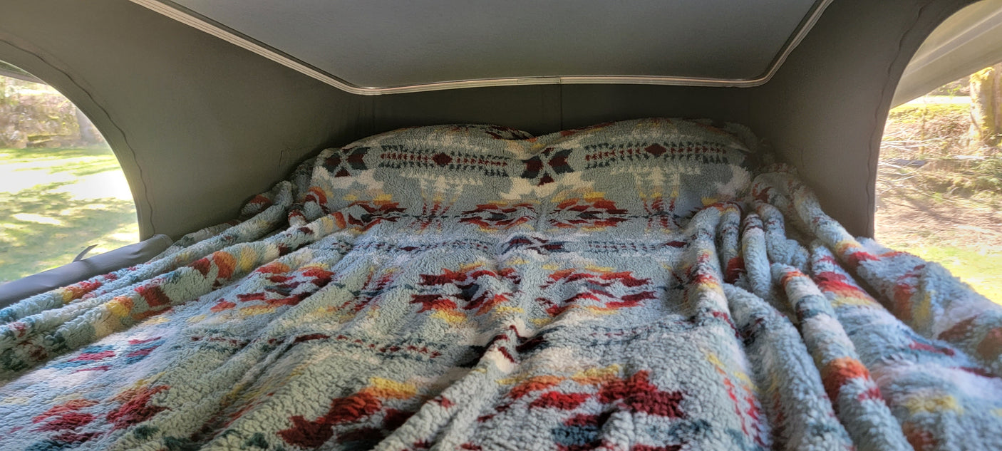 Inside an RV rental: a light blue, fluffy patterned blanket on a cozy pop-top bed, with outdoor views.