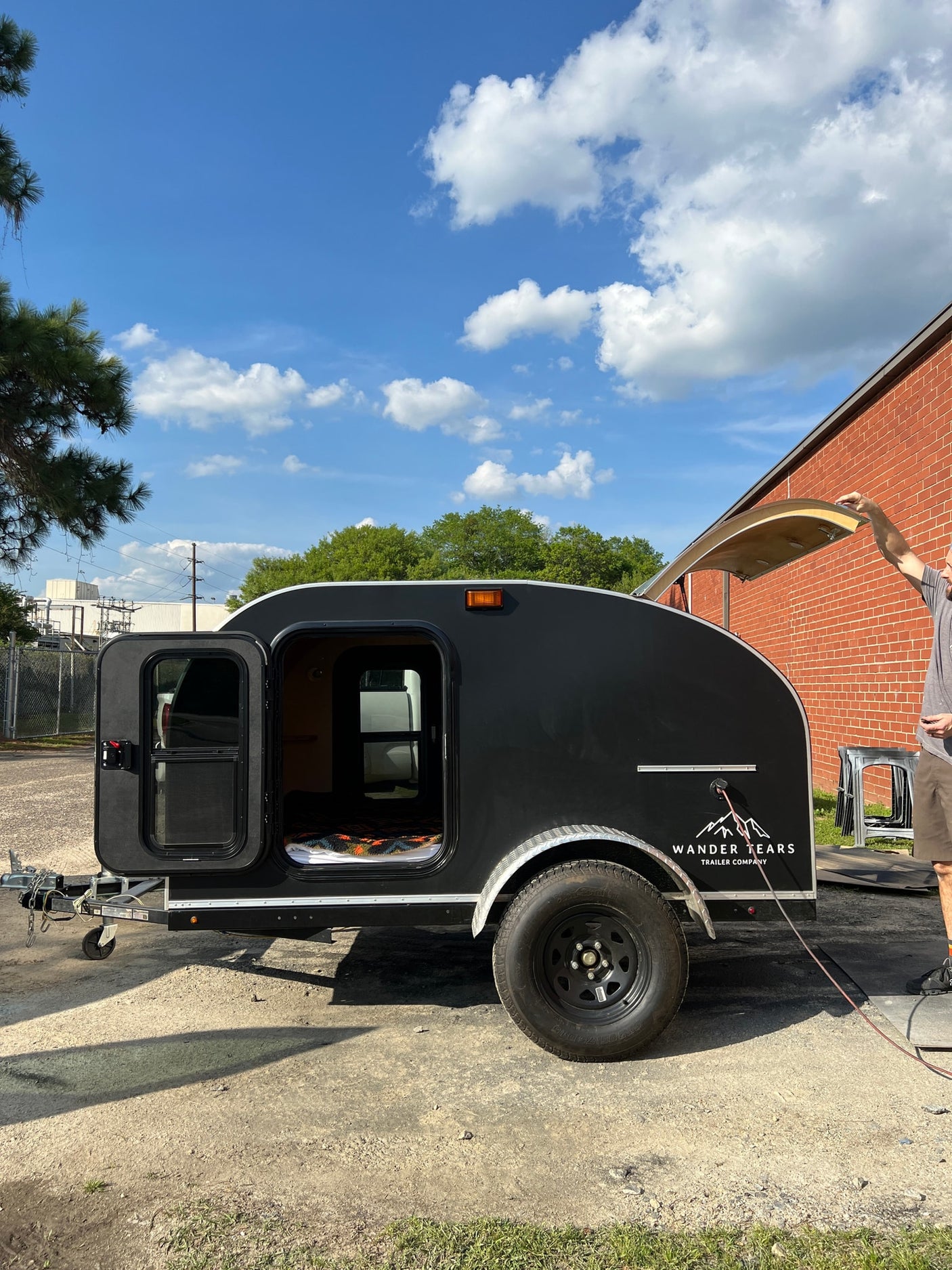 showcasing wooden storage and black exterior with silver trim.|Man at a serene forest campsite with his teardrop camper