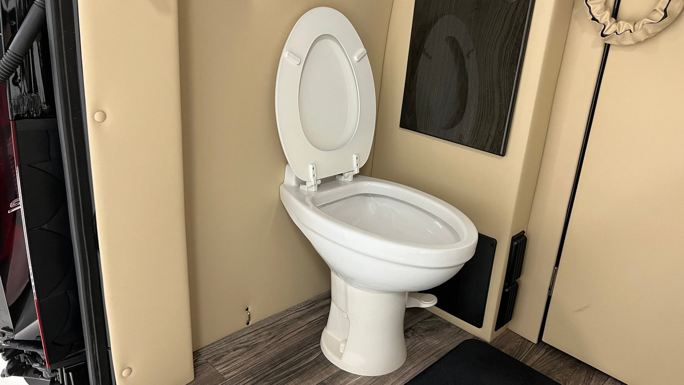A clean white toilet with an open lid in an RV rental bathroom. Features beige walls, wood-look floor, and dark accents.