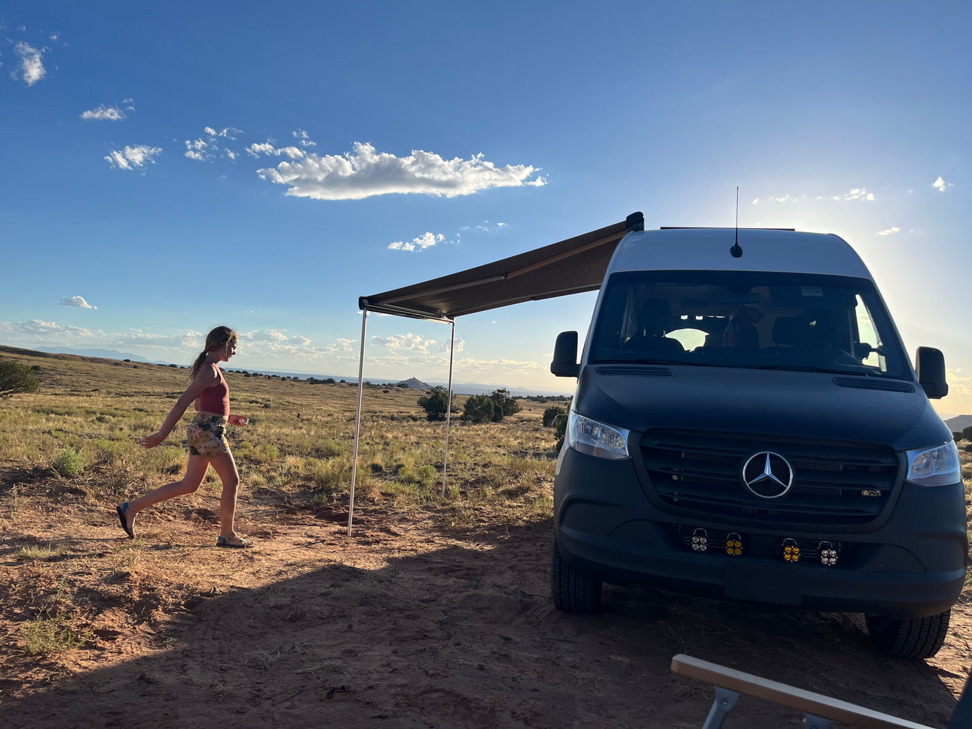 RV rental adventure: A woman walks by a camper van with an awning in a sunny, expansive desert landscape.