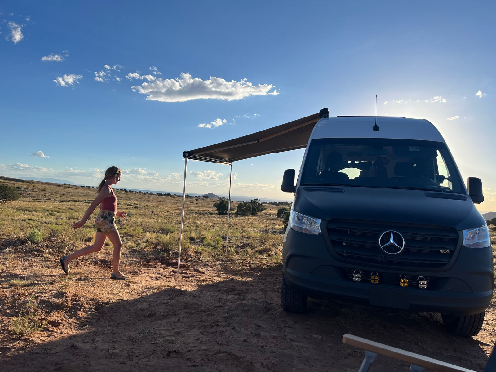 RV rental adventure: A woman walks by a camper van with an awning in a sunny, expansive desert landscape.