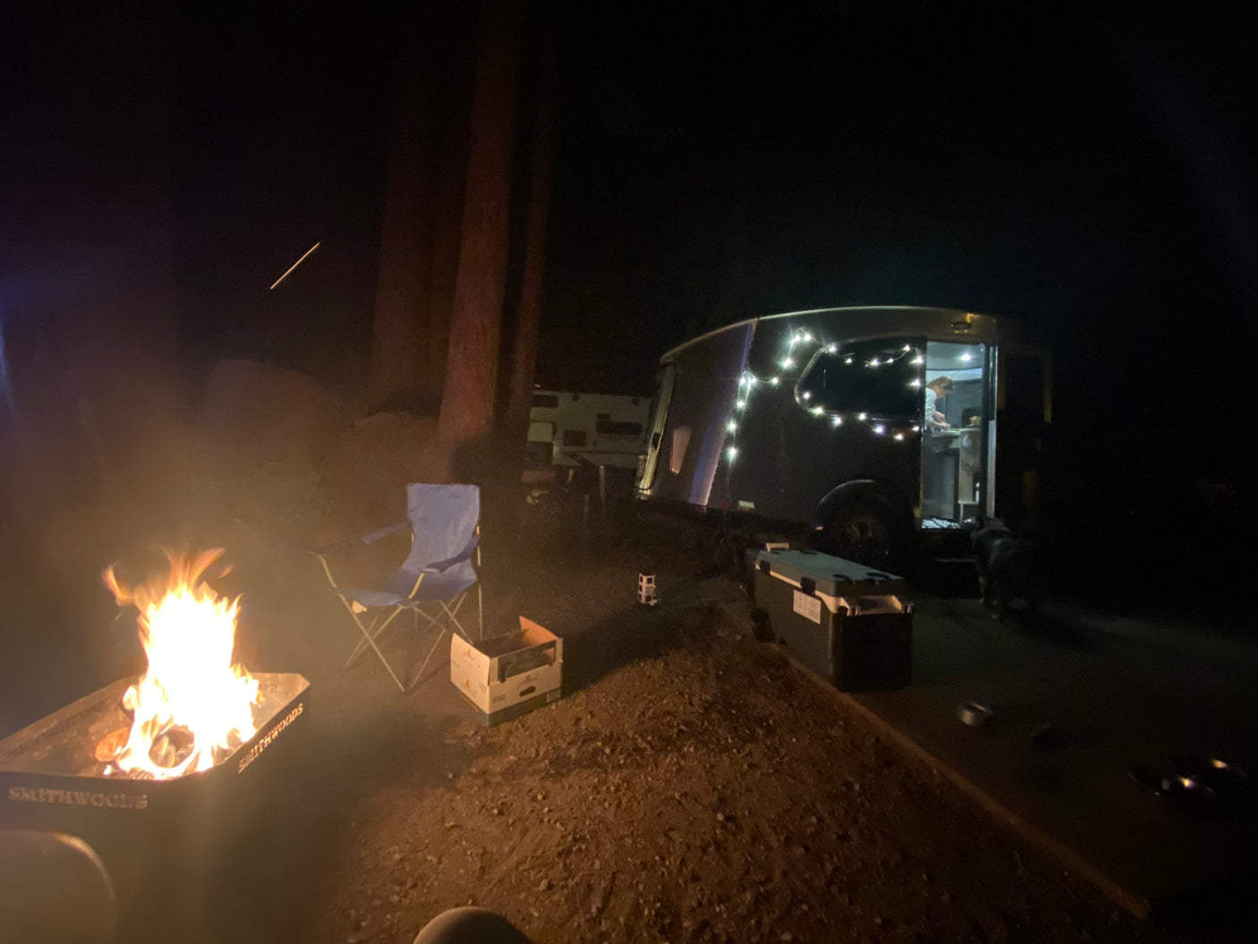 Nighttime RV rental scene: bright campfire, person inside lit trailer, dog, & camping chair for outdoor relaxation.