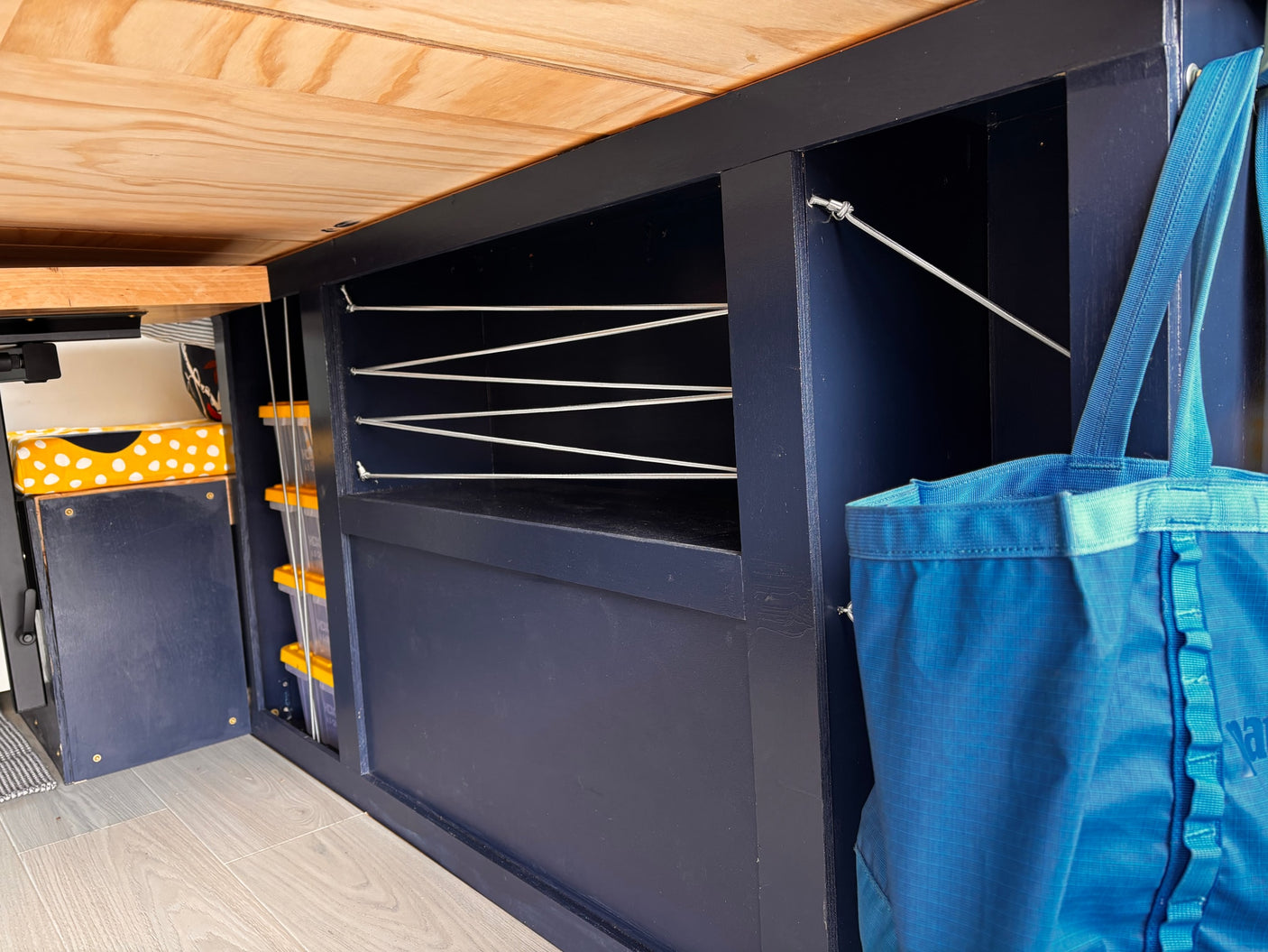RV rental interior featuring custom dark blue cabinet storage with elastic cords, securing items like clear containers. A blue tote bag hangs nearby, and the ceiling is light wood.