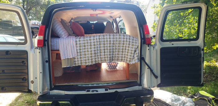 Converted van RV rental interior: cozy bed with pillows, patterned bedding, & under-bed storage, with open rear doors.