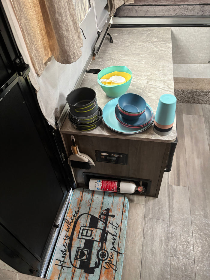 RV rental kitchen: camping dinnerware, pots, cups, and mixing bowls on the counter. Fire extinguisher and "Home is where you park it" mat.