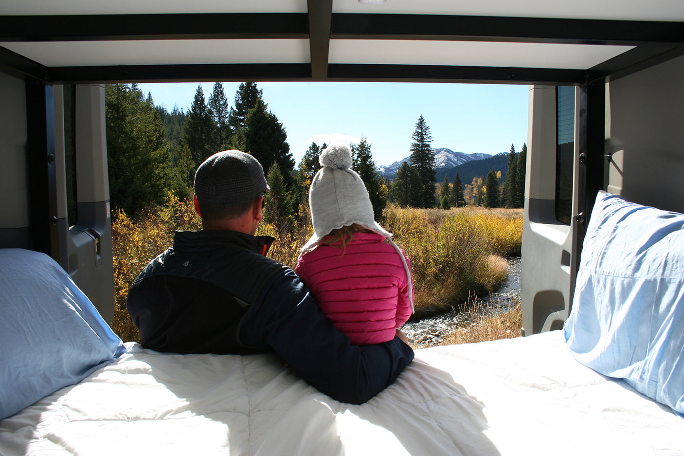 Inside their RV rental, a father and child gaze at a stunning mountain, forest, and river view. Perfect road trip stop.