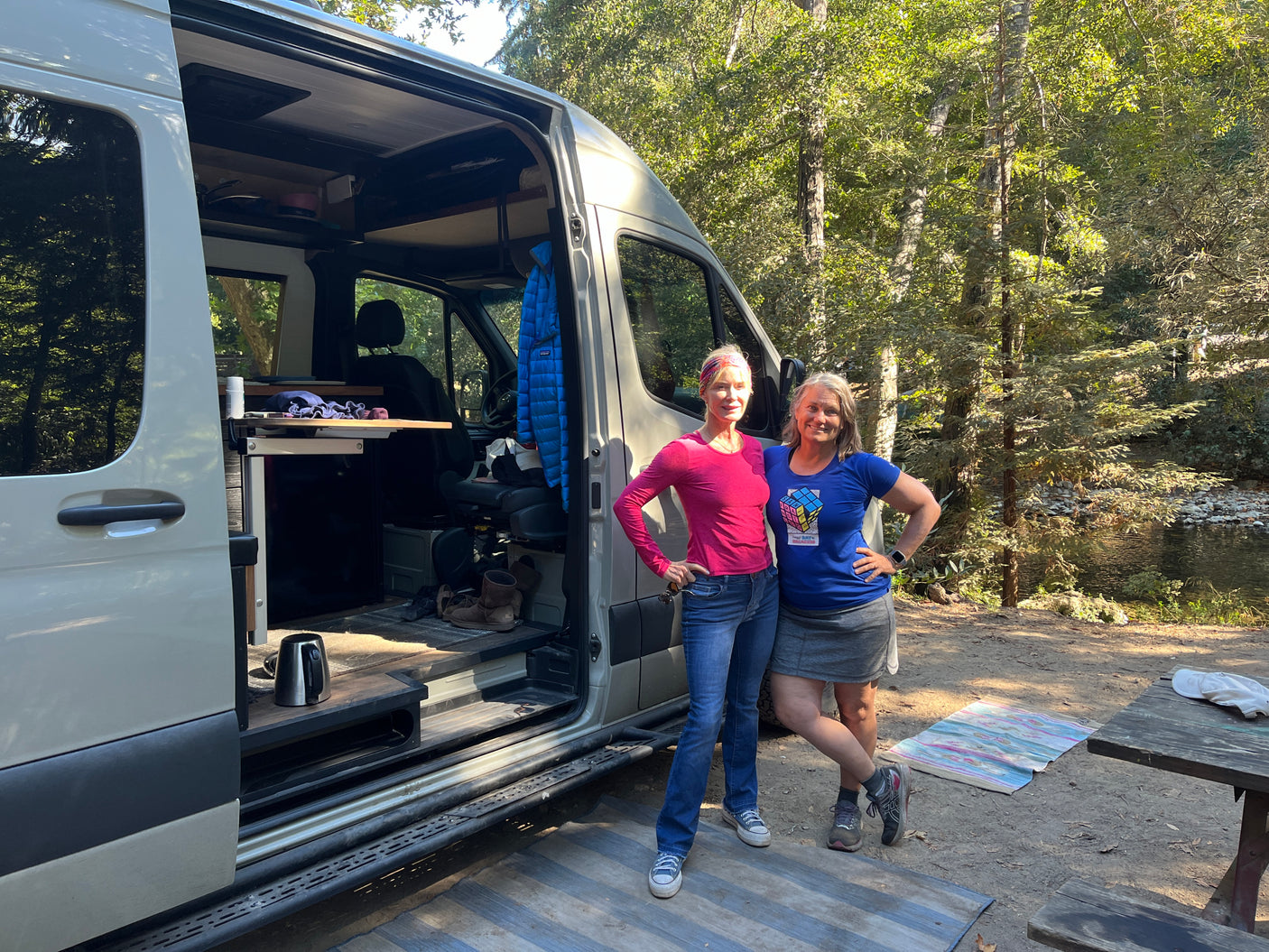 Two friends beside their custom rv rental van, open to show interior. By a river in a wooded campsite.