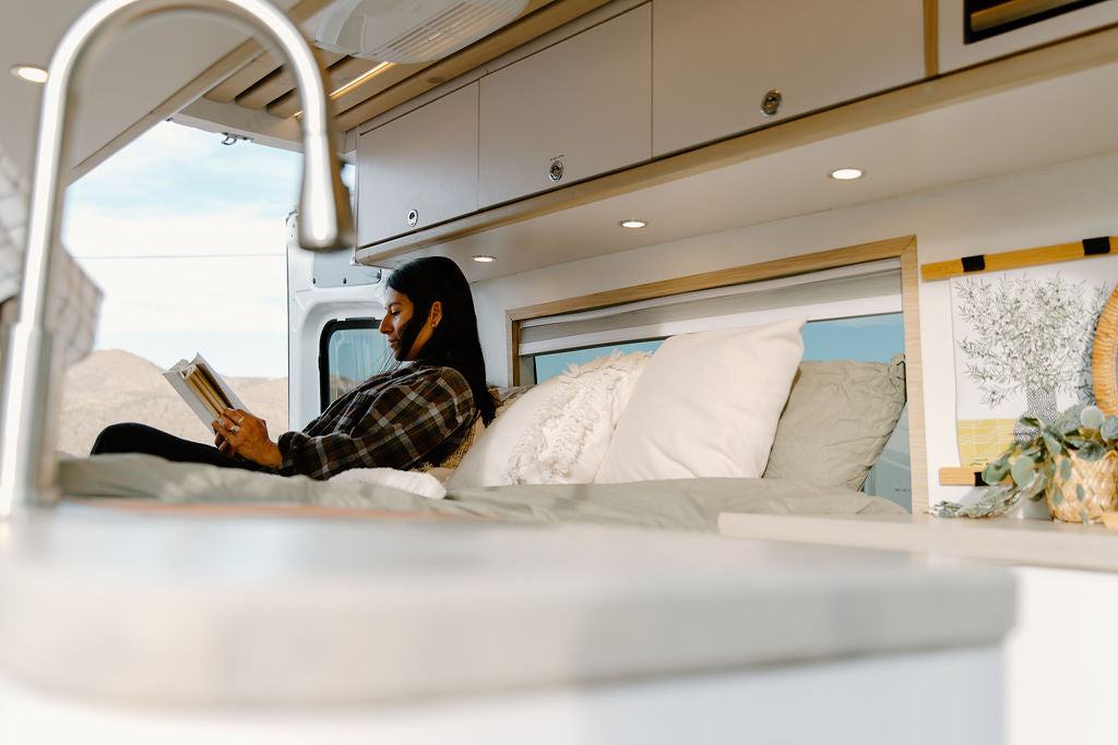 Relax in an RV rental! A woman reads a book comfortably on a bed inside a modern camper van with a scenic view.