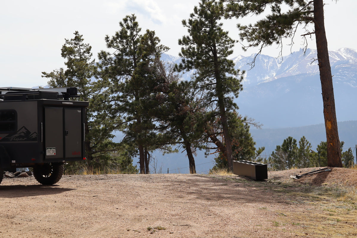 Adventure awaits! A dark gray RV rental trailer sits on a dirt road surrounded by pine trees and distant snow-capped mountains. — photo 18