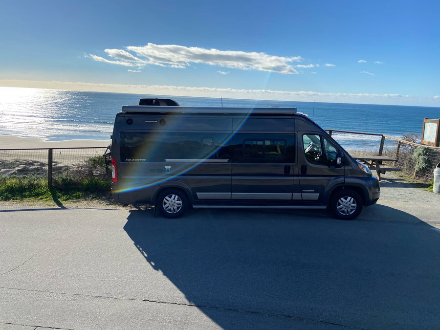 Enjoy an RV rental: a dark grey camper van parked by a sunny beach and ocean view. Ideal for road trip adventures!