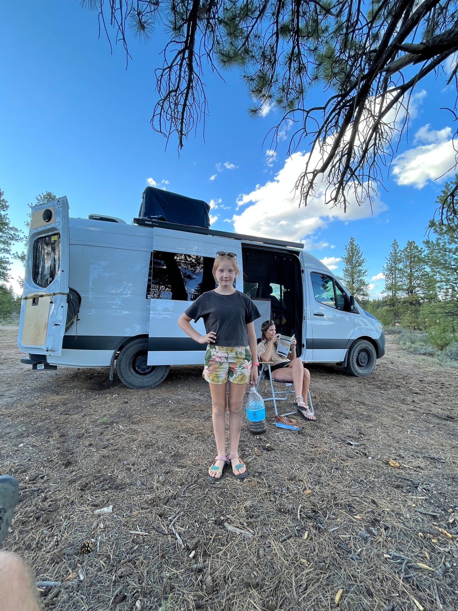 Relaxing van life with an RV rental. A girl stands while an adult reads by their white camper van in nature.