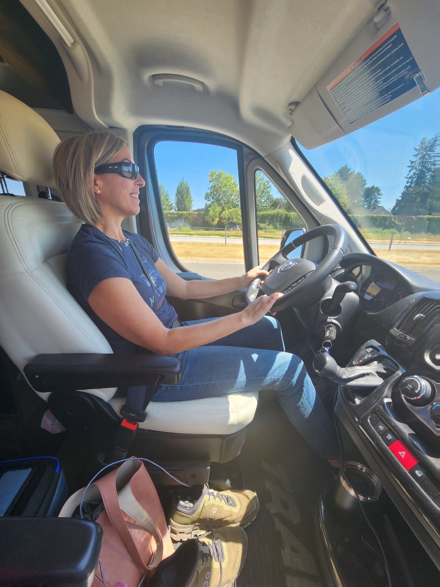Woman in pilot seat of an RV rental van, smiling and driving. Bright interior shot during a sunny road trip.