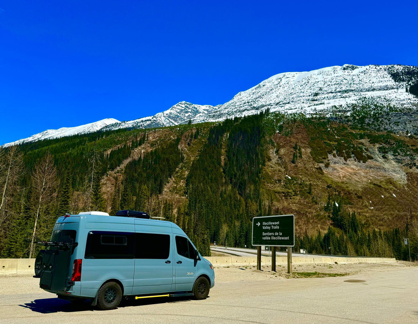 Light blue **rv rental** van at base of snowy mountains & forest. Sign for Illecillewaet Valley Trails. Scenic road trip adventure.