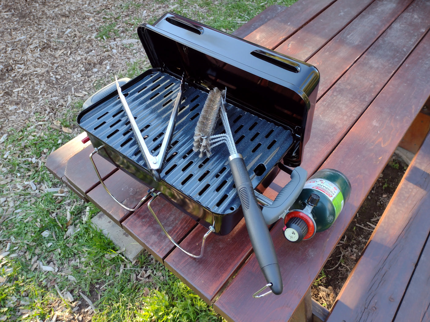 Compact propane grill, tools, and Coleman tank ready for RV rental outdoor cooking on a picnic table.