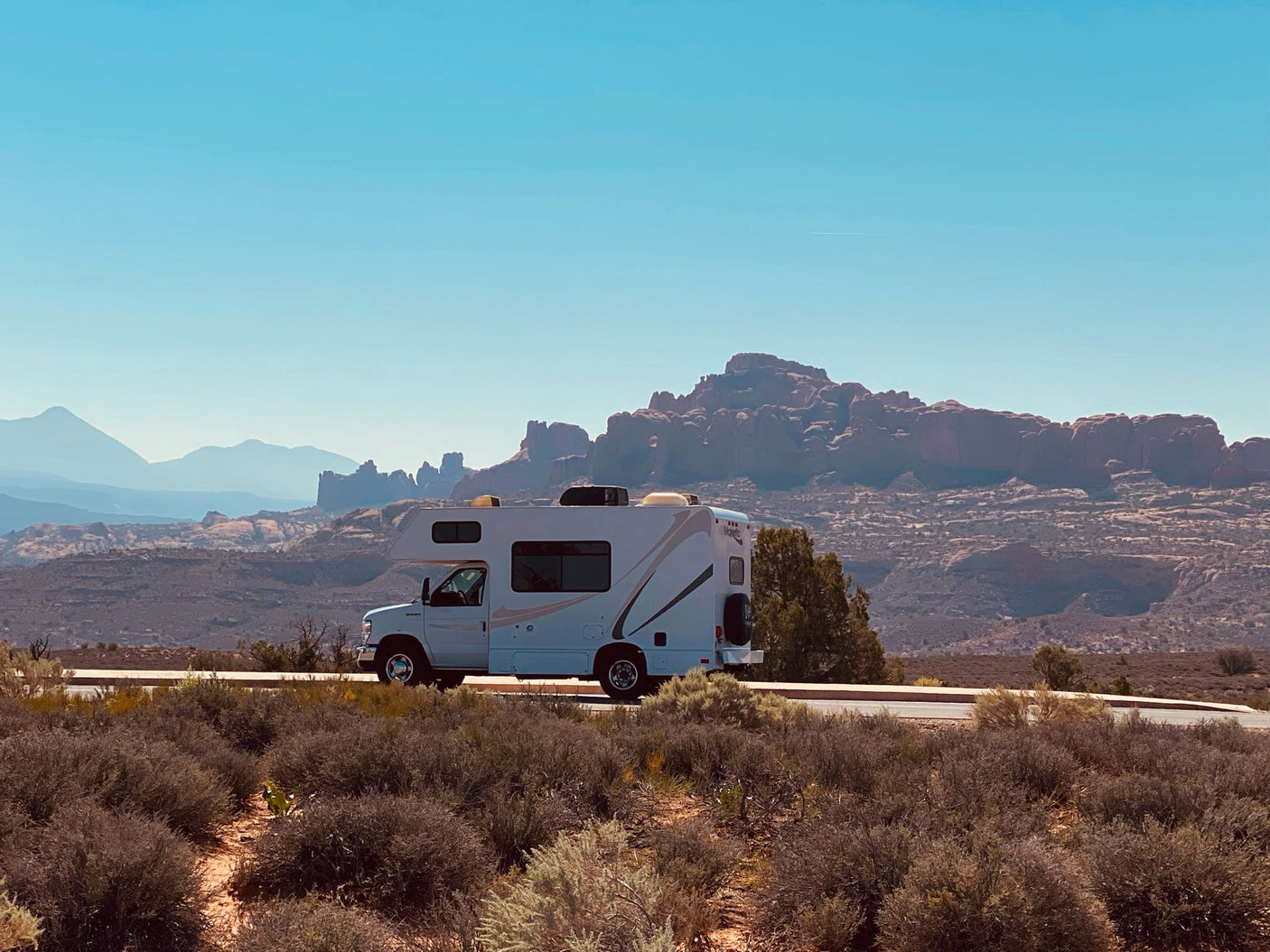 White RV rental parked on a scenic desert road, with distant red rock mountains layered against a clear blue sky. Perfect for a road trip.